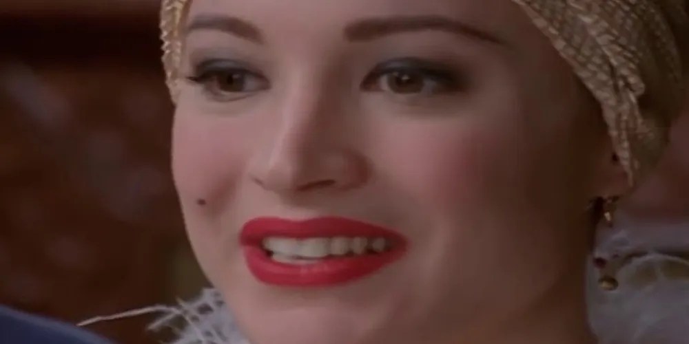 Close up expression of Madame Visage speaking, with pink lipstick, lightly powdered face, golden earrings, and fancy head wrap.