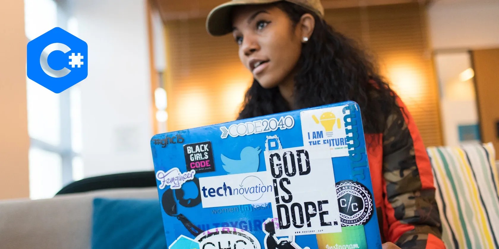 Woman sitting on a sofa, holding a blue laptop covered in stickers, facing toward the camera