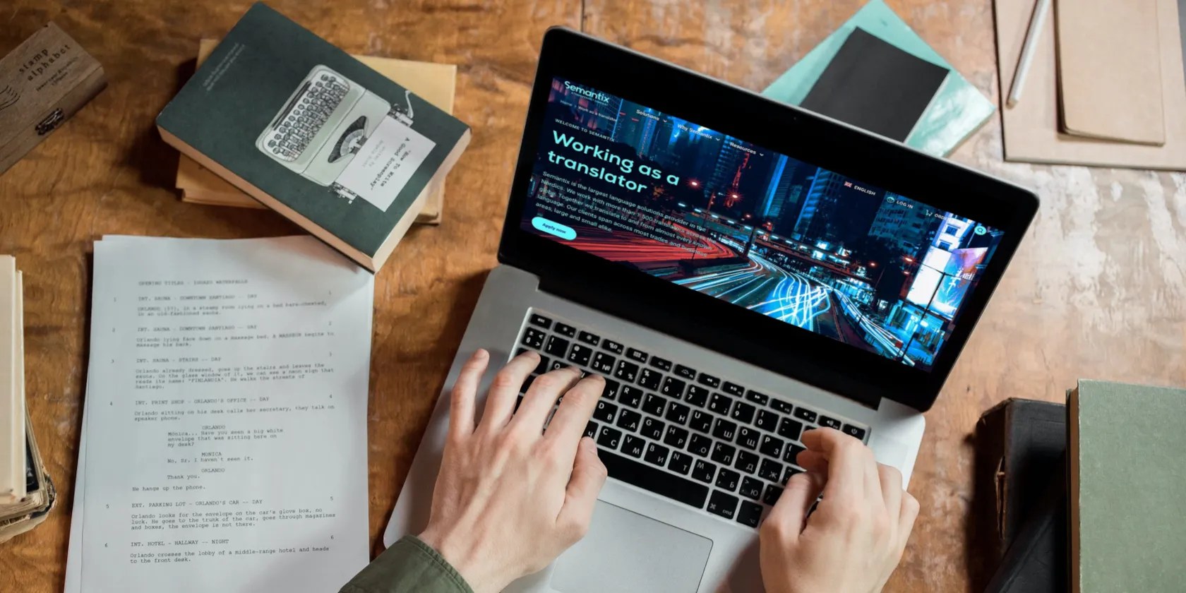 Person working on a MacBook viewing a Translation Works website surrounded by books and papers