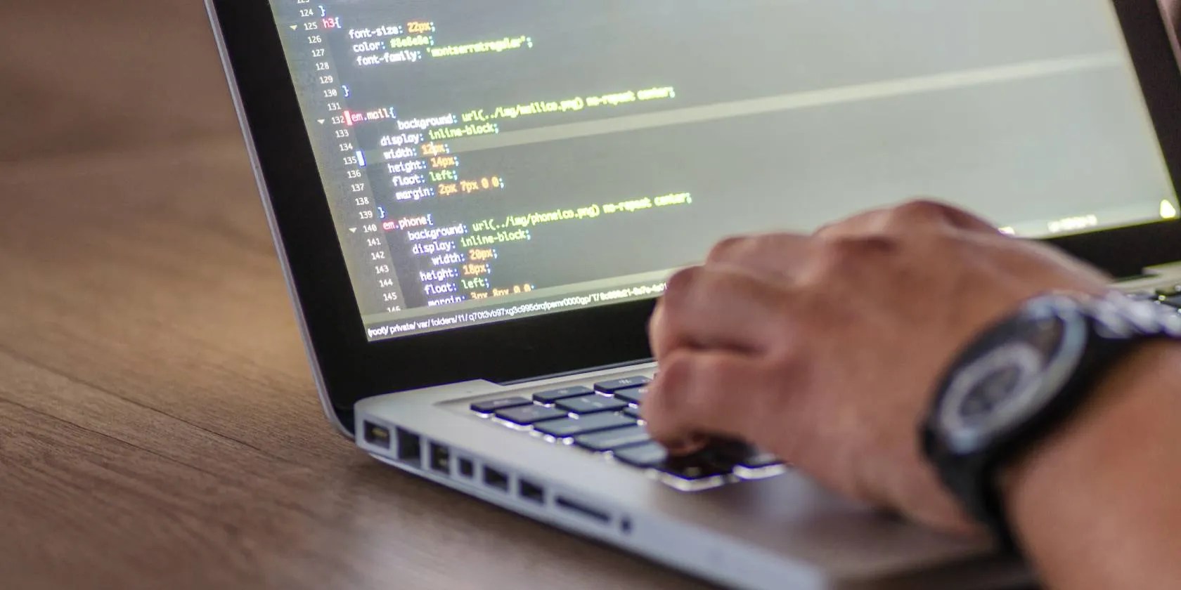 A close up of a hand typing on a laptop keyboard. The screen shows some CSS code.