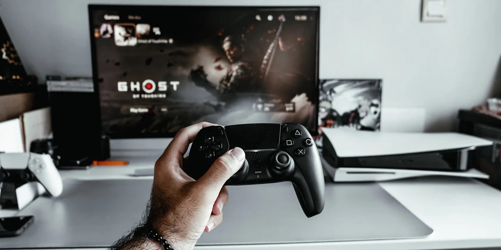 A hand holding a DualSense controller with a PS5 and monitor in the background