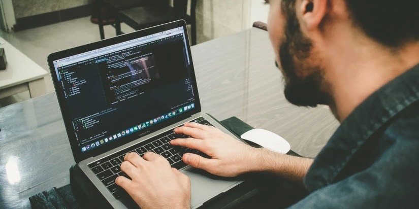 A man sitting at a desk typing on a laptop with code on the screen.