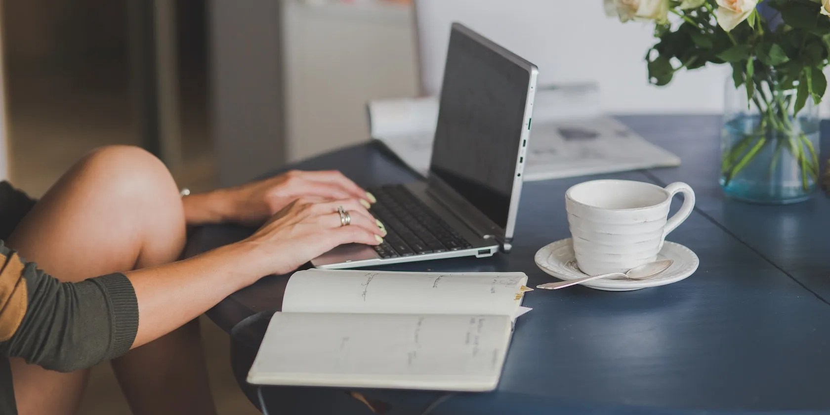 Someone typing casually on a laptop next to an open diary and an empty teacup.