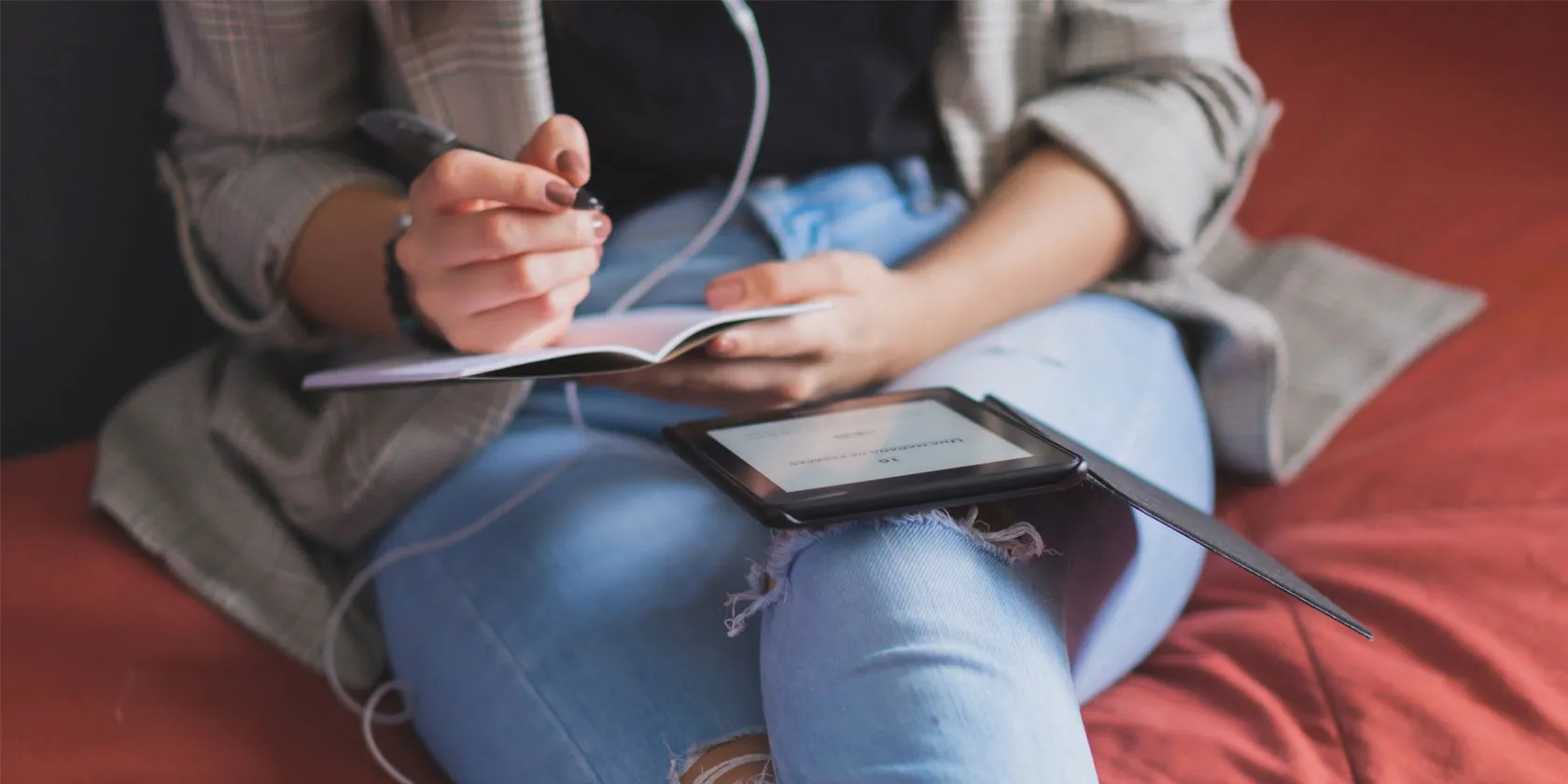 Close-up of woman’s lap as she writes in a notebook while viewing an e-reader