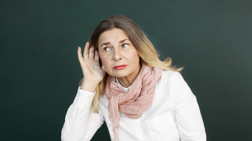 Studio shot of stylishly dressed beautiful retired female with blonde hair holding hand at her ear while overhearing interesting conversation, struggling to hear some news, secret or gossip