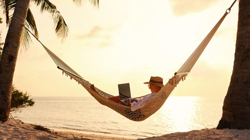 Woman in a hammock on the beach working on a laptop.