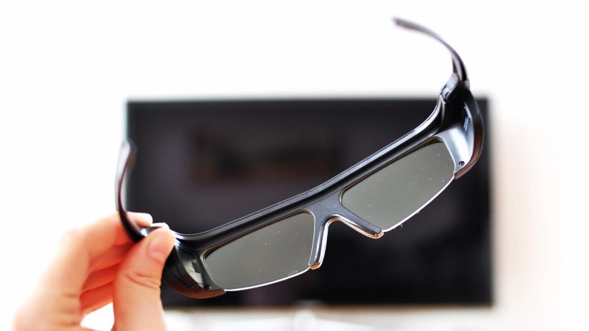 Person&rsquo;s hand holding 3D glasses in front of a TV.