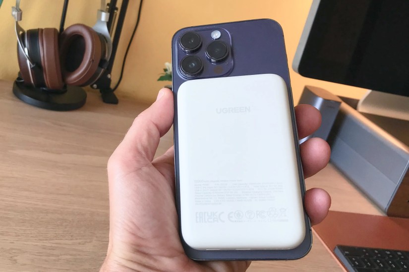 An iPhone held in hand over a wooden desk with a Ugreen MagSafe power bank snapped onto the back.