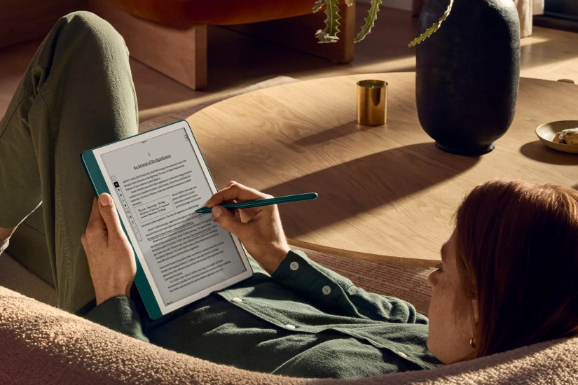Woman lying on a couch, using Amazon&rsquo;s Kindle Scribble reader with a stylus to add inline notes.