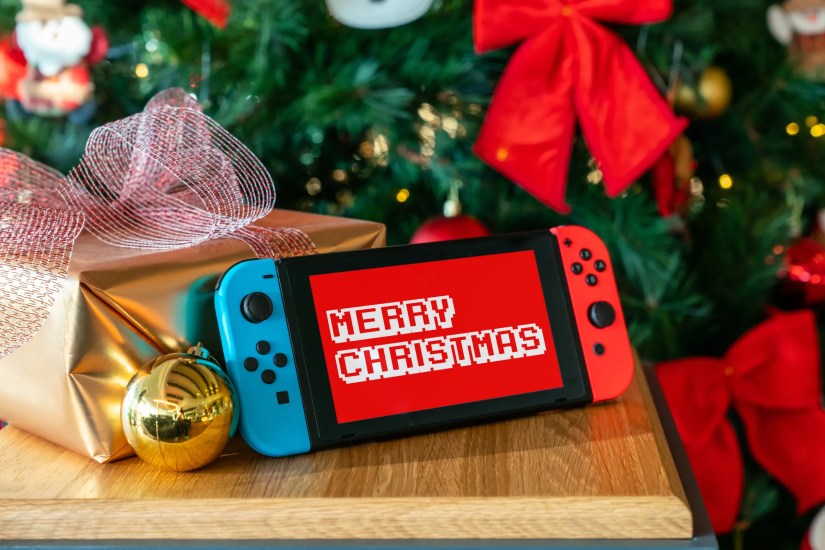 Nintendo Switch with &lsquo;Merry Christmas&rsquo; on the screen sat in front of a Christmas tree.