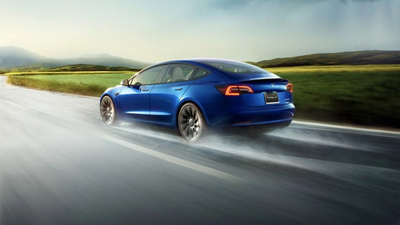 A blue Tesla Model 3 driving on a country road with surface spray coming off the road.