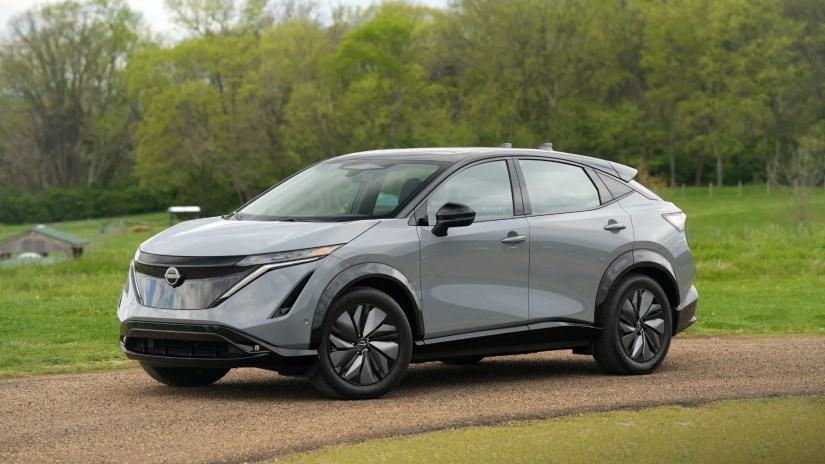 Front 3/4 view of a gray 2025 Nissan Ariya parked on a gravel track with grass and trees in the background.