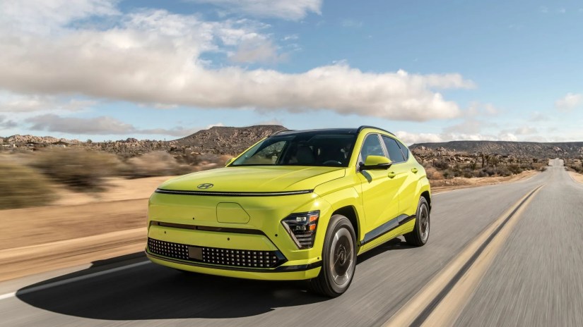 Front 3/4 view of a yellow 2025 Hyundai Kona Electric driving on a road through the desert.