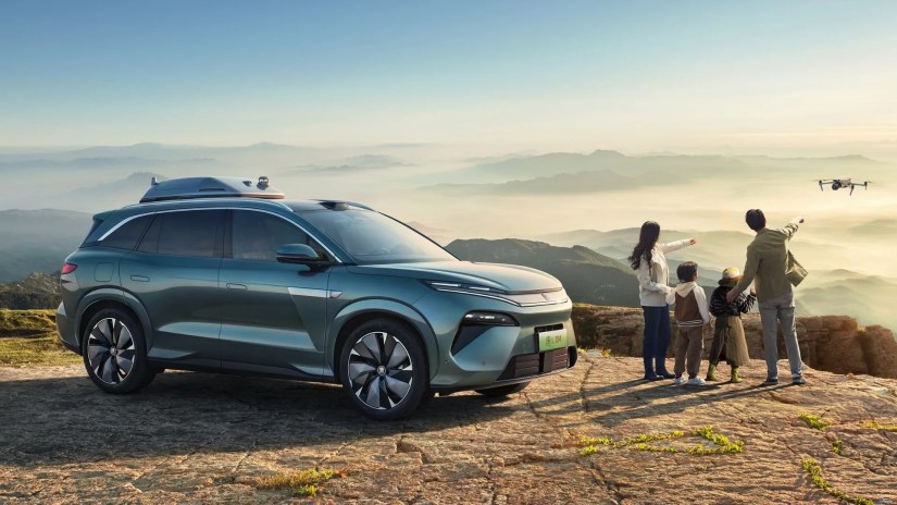 Front three-quarter view of the BYD Tang L with a family stood beside it looking out at the distance as a drone flies overhead.