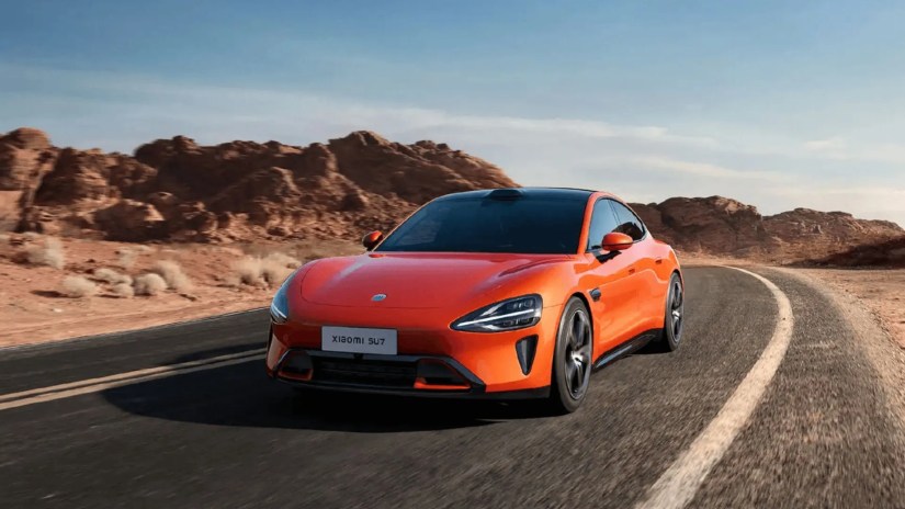 Front 3/4 view of an orange Xiaomi SU7 driving on a winding road through the desert.
