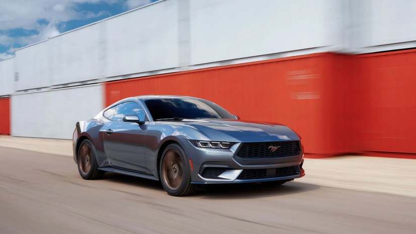 Front 3/4 shot of a gray 2024 Ford Mustang driving past shipping containers.