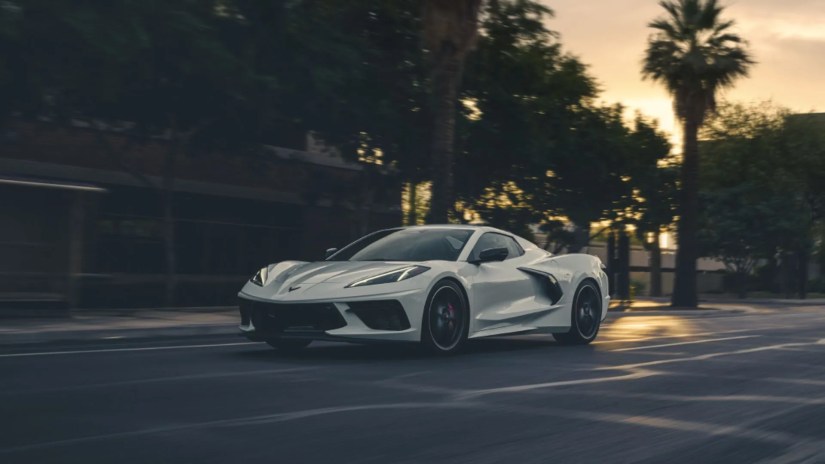 Frotn 3/4 shot of a white 2025 Corvette Stingray driving through a city as the sun sets.