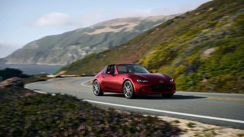 24MY-MX-5-8Front 3/4 shot of a red 2024 Mazda MX-5 Miata RF driving oin a winding road with the ocean in the background.