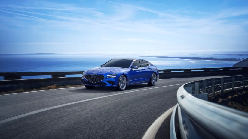 Frotn 3/4 shot of a blue 2025 Genesis G70 driving on a winding road beside the ocean.