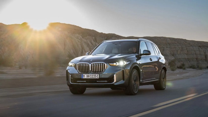 Dynamic front 3/4 shot of a blue 2024 BMW X5 xDrive50e driving on a country road as the sun sets.