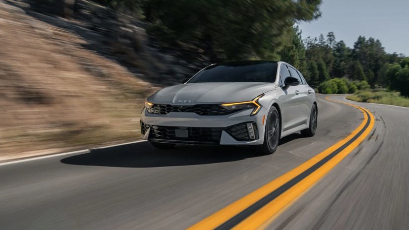 Dynamic front 3/4 shot of a gray 2025 Kia K5 driving on a country road through the mountains.