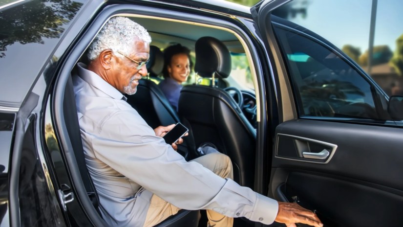 An older gentleman getting out of the backseat of a car while a woman looks back at him smiling.