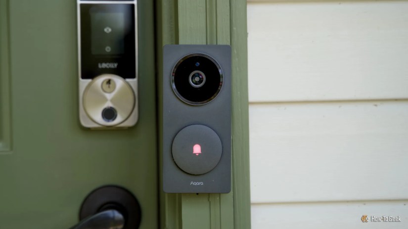 A head-on shot of the Aqara Doorbell Camera Hub G410 mounted to a green door frame with cream house siding next to it.