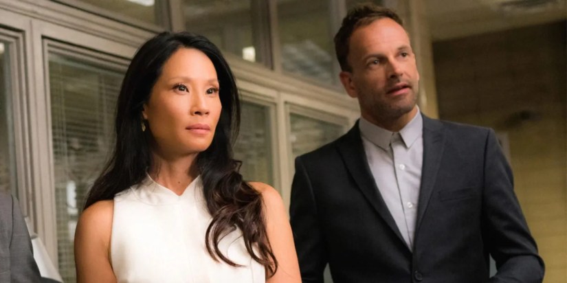 Holmes (Jonny Lee Miller) and Watson (Lucy Liu) Look On in Elementary