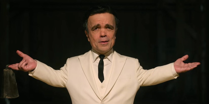 Peter Dinklage is walking out of the shadows with his arms out to the side.