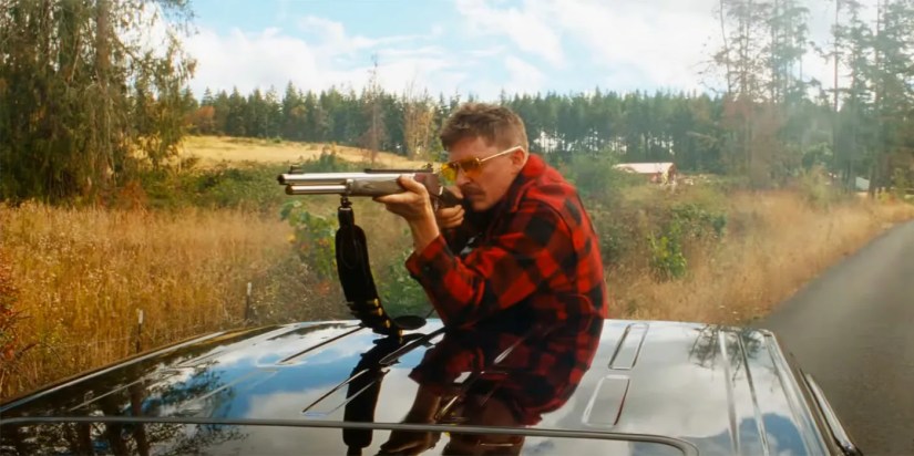 Kyle Gallner perched at the top of a moving car as he aims his gun in Strange Darling