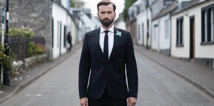 David Tennant as Tom Kendrick wearing a black suit and standing in the middle of the street in Deadwater Fell.