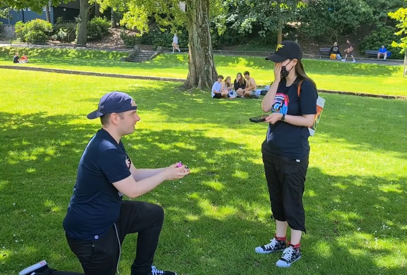 A man on one knee in the park holding a Love Ball proposing to a woman who looks surprised.