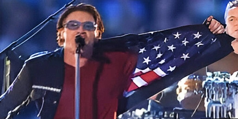 Bono is in the middle of the screen shown from his chest up. He has on sunglasses, a maroon shirt, and he holds open the left side of his jacket to show the American Flag inside. Behind him is the drummer to the right, and a microphone stand to the left. Everything else is fuzzy blue.