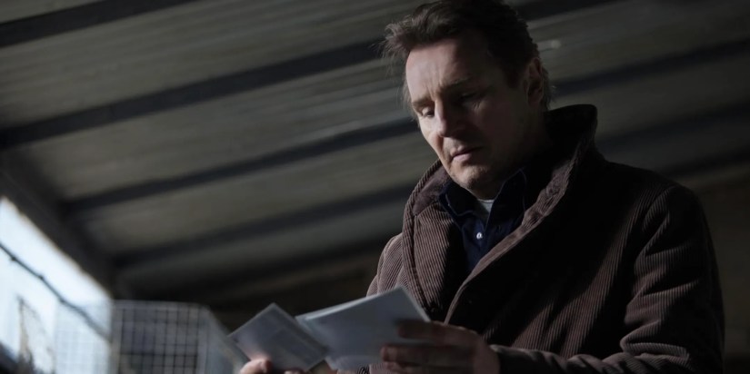 Liam Neeson looks over a handful of cards in his hands.