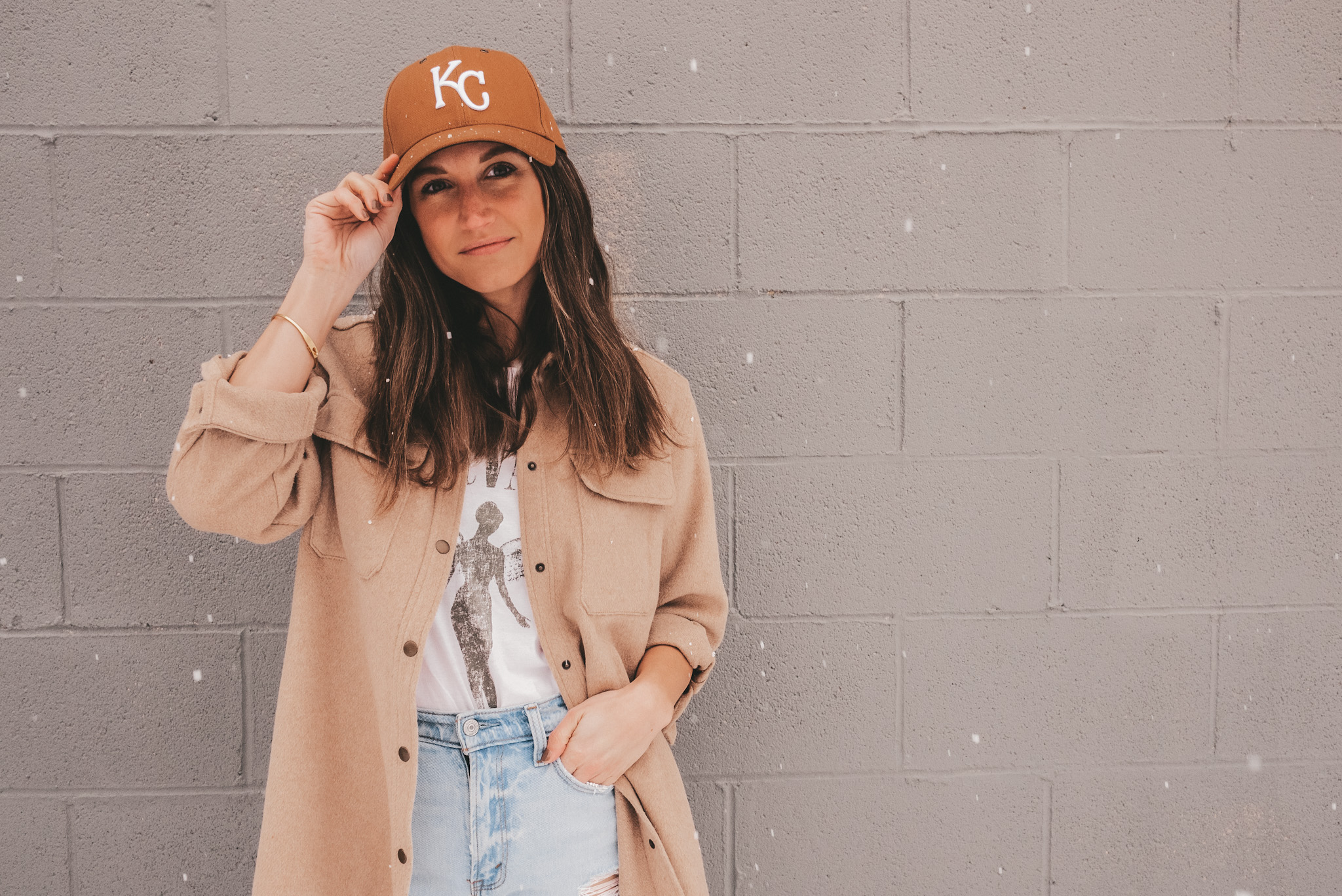 ashley standing on a snowy sidewalk wearing a brown KC baseball hat, a tan colored shacket, a white graphic tee, light washed distressed jeans and white sneakers with a gray brick background