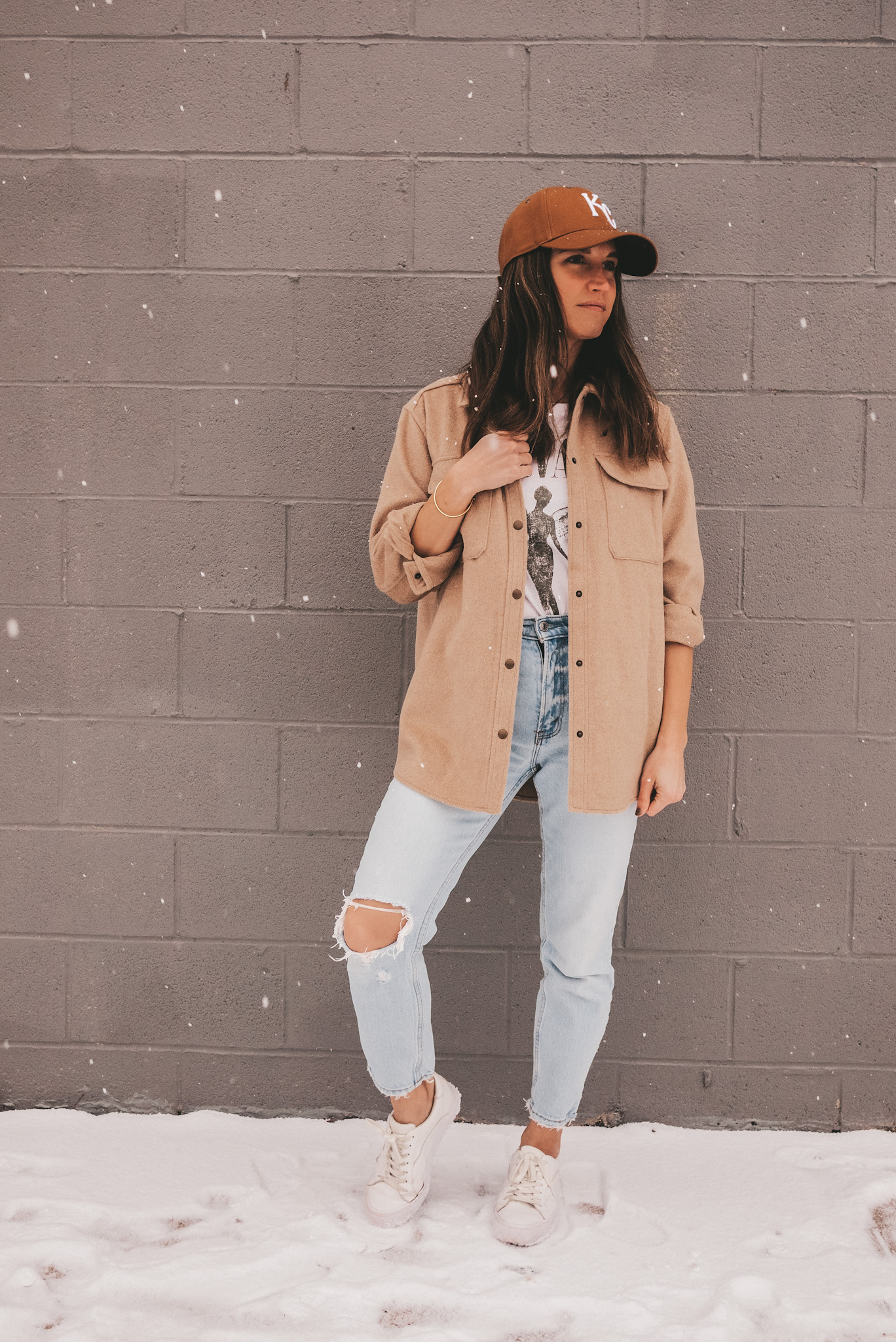 ashley standing on a snowy sidewalk wearing a brown KC baseball hat, a tan colored shacket, a white graphic tee, light washed distressed jeans and white sneakers with a gray brick background