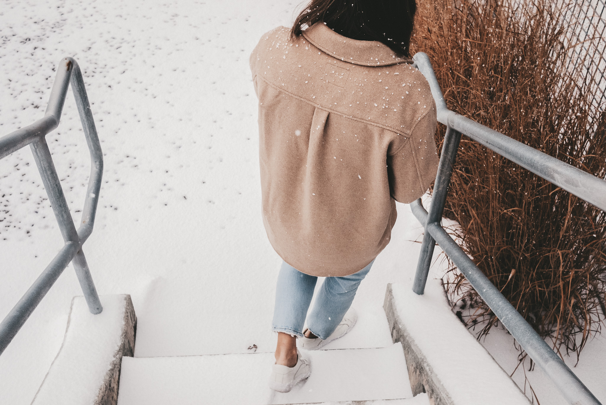 ashley walking down snowy stairs wearing white sneakers, light wash jeans and a tan colored shacket