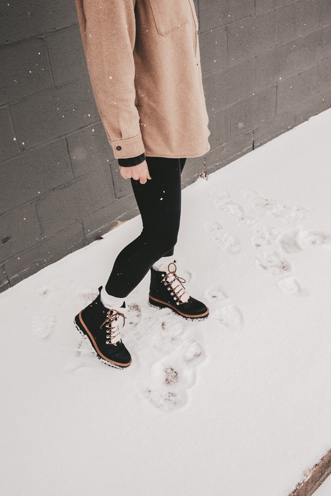 Ashley wearing a tan colored shacket, Spanx faux leather leggings, black winter boots with faux sherpa lining standing in the snow with a gray brick background