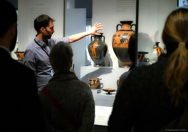 ExL educator Stephen Guerriero stands in front of and gestures toward two black and orange ancient grecian urns during a discussion at the Museum of Fine Arts in Boston