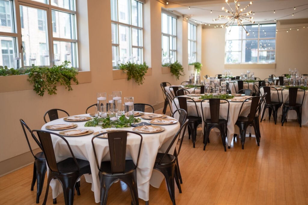 Round reception tables are scattered in a brightly-lit space with greenery on the windowsills and string lights hanging above
