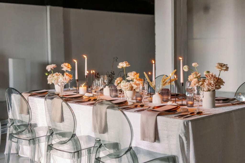 Clear chairs and neutral linens decorate a reception table with small vases of light-colored flowers next to tall taper candles