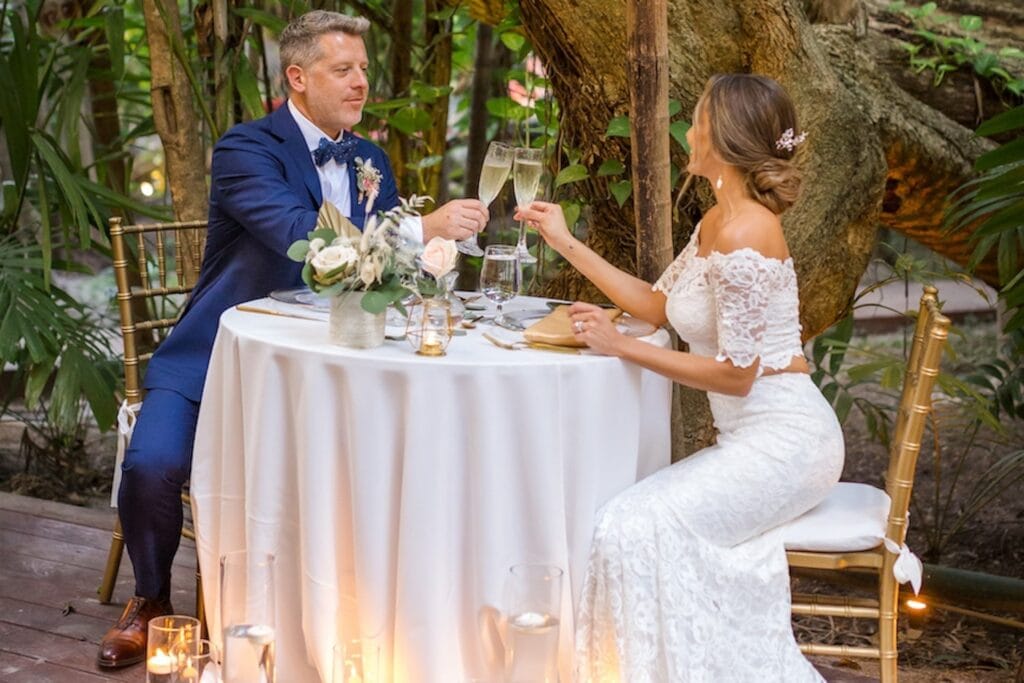 A bride and groom toast each other in gold chairs at a small round table set among palm trees