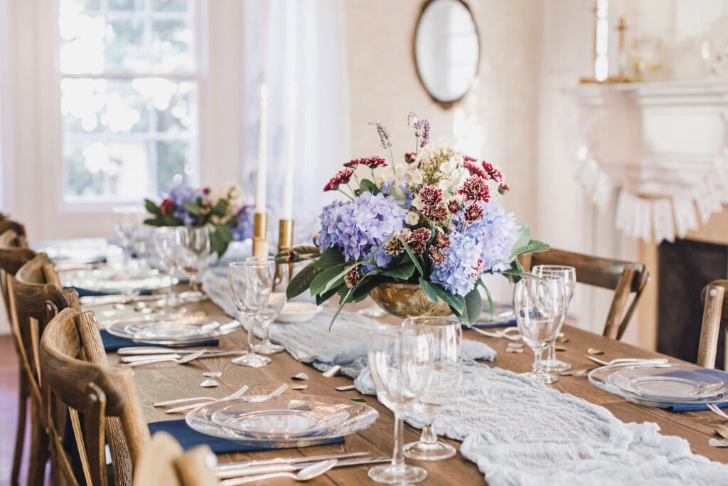 Purple hydrangea in gold vase on top of a wooden table in a bright, cheery dining room set up for an intimate dinner