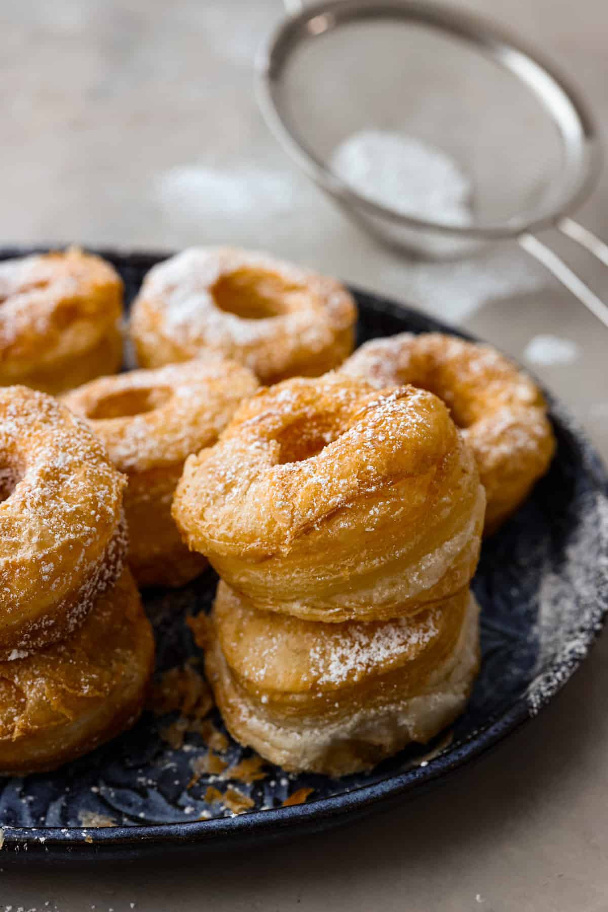 Get ready to discover the joy of homemade cronuts with this delicious recipe! Perfectly flakey and fried, this easy-to-make recipe is perfect for any occasion. A platter of cronuts sprinkled with powdered sugar.