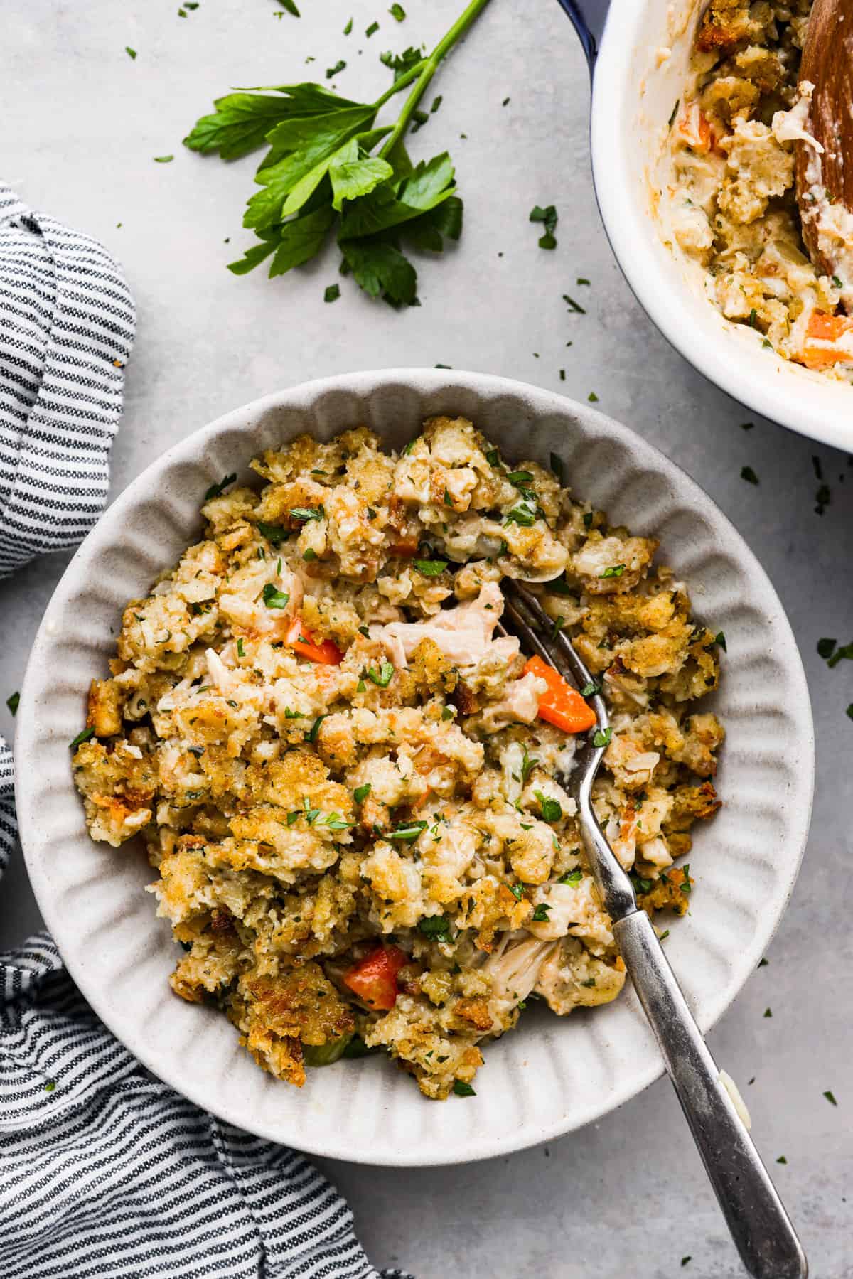 A delicious, hearty casserole made with stuffing mix, shredded chicken, vegetables, and all the tasty seasonings. Chicken and stuffing casserole is a fun twist on a classic dish and the perfect addition to your Thanksgiving lineup! A serving of casserole in a white scalloped bowl.