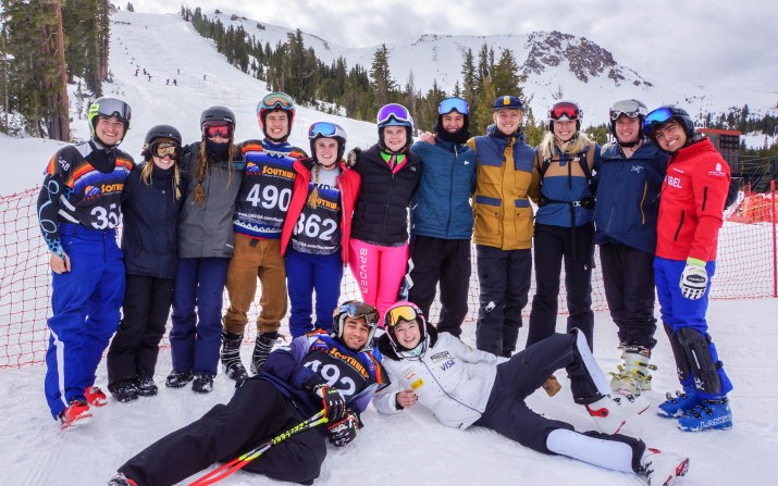 Thirteen students, eleven standing and two laying on the ground, wear ski coats, pants, and goggles with four wearing competition jerseys.