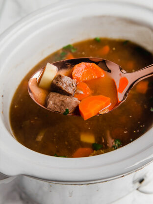 A slow cooker filled with beef stew and a serving spoon taking some out of it.