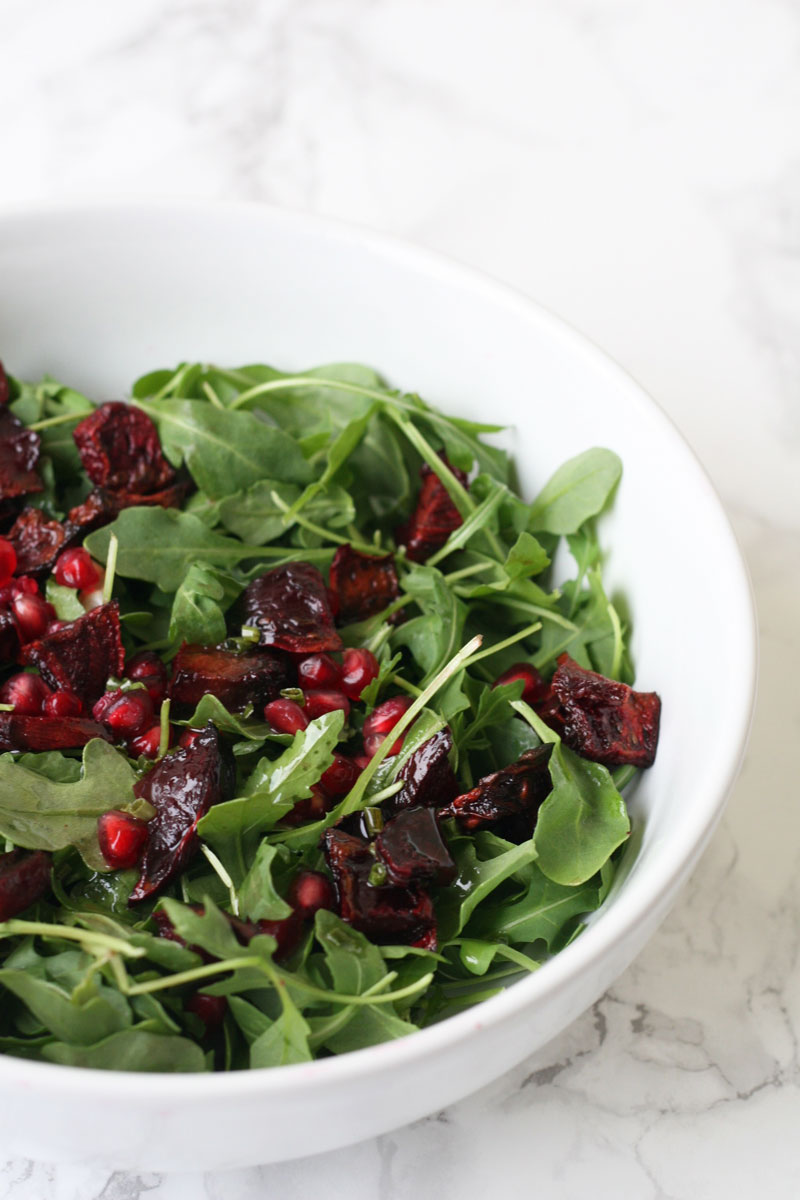 Arugula salad in a white bowl with pomegranate seeds and roasted beets.