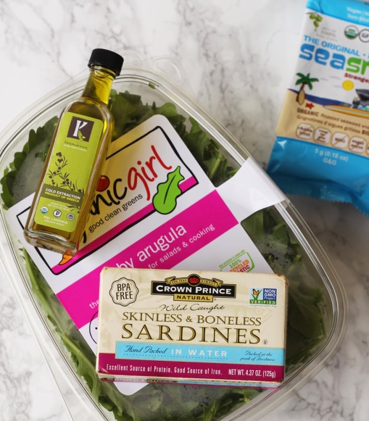 A container of arugular, olive oil, sardines, and seaweed snacks out on a countertop.