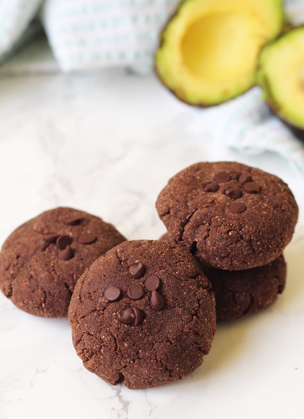 A pile of avocado cookies on a counter with avocado behind it.